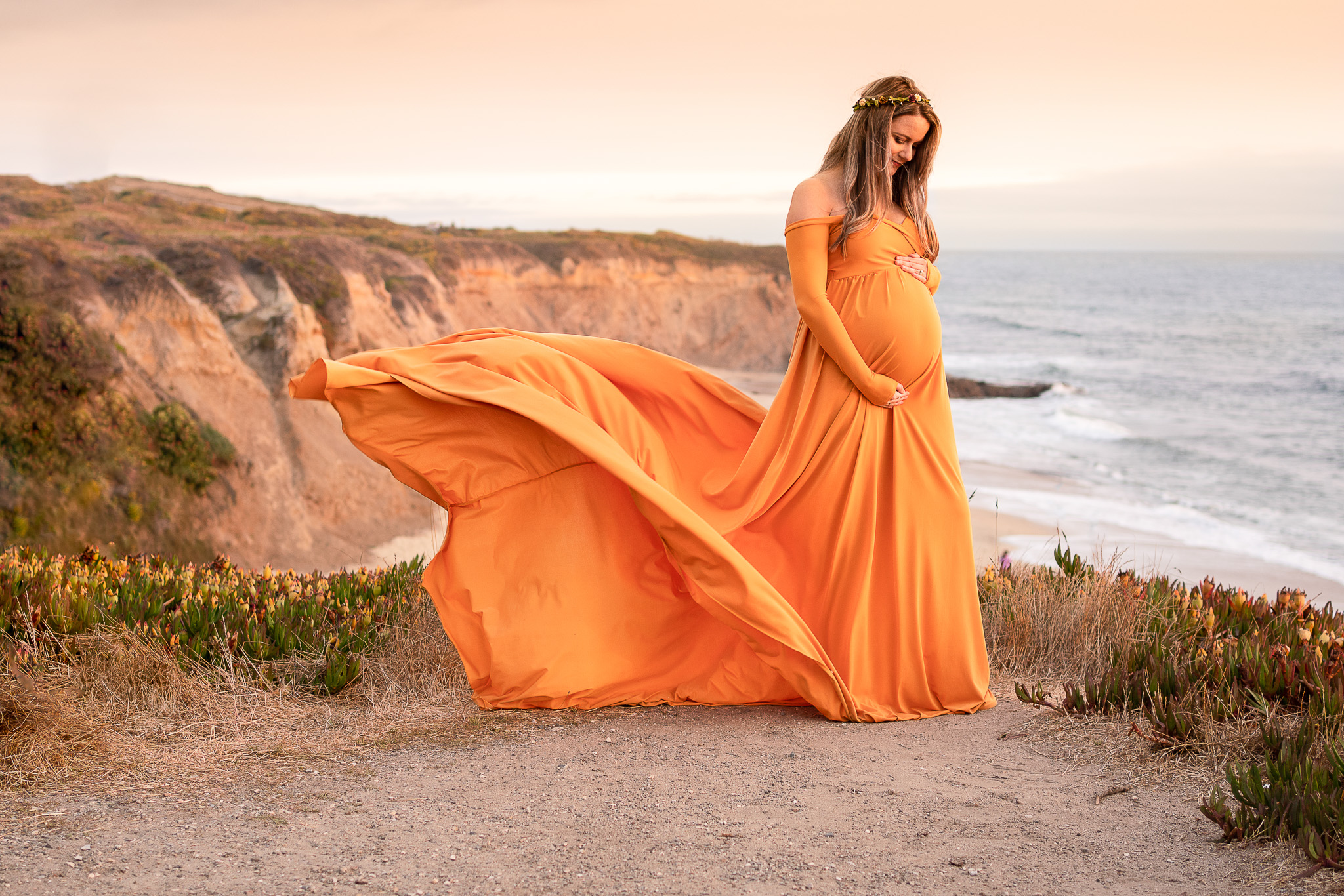 Pregnant woman in a long orange dress with backdrop of cliffs and ocean behind her in half moon bay