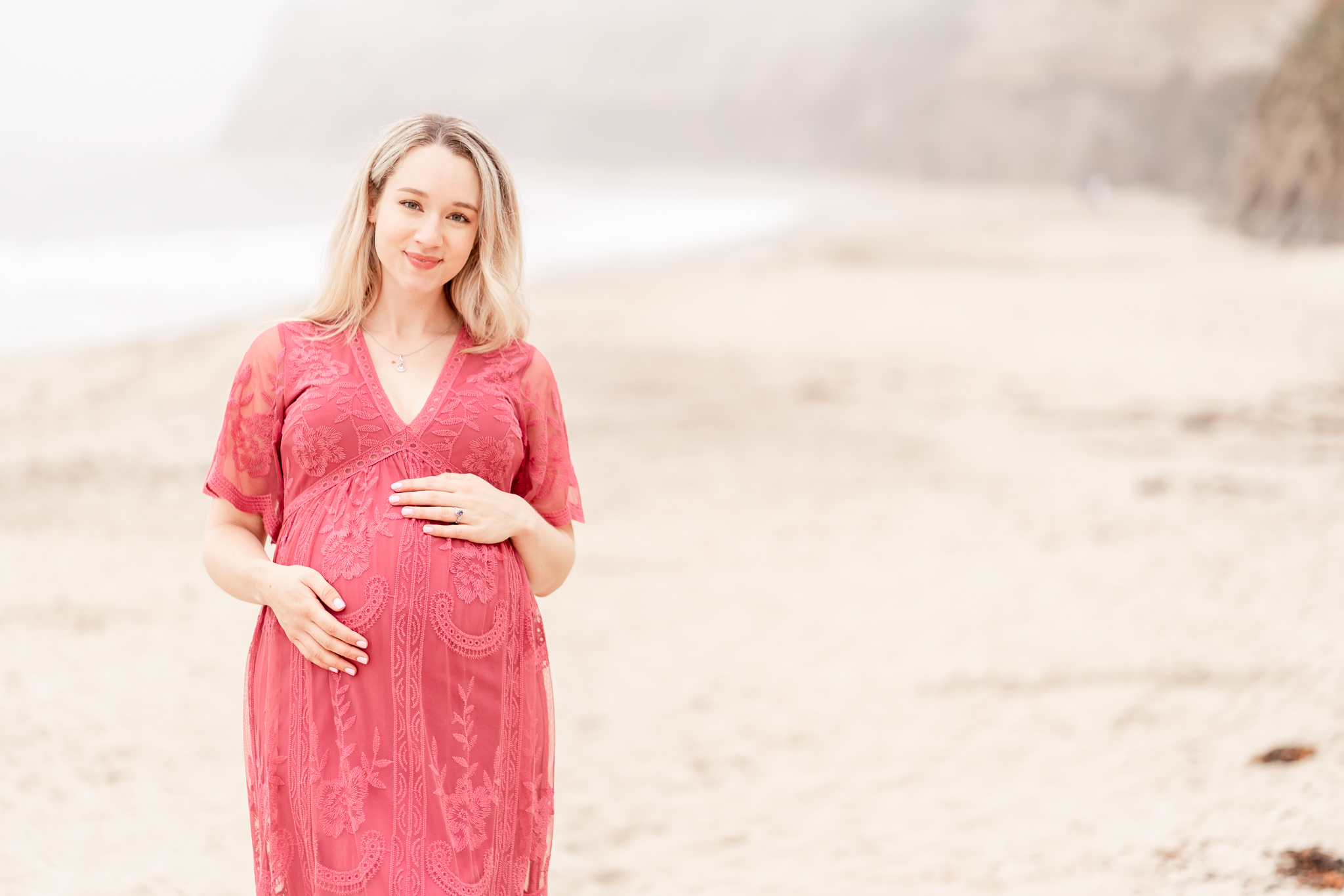 Pregnant woman standing on a beach wearing a pink dress and smiling at the camera
