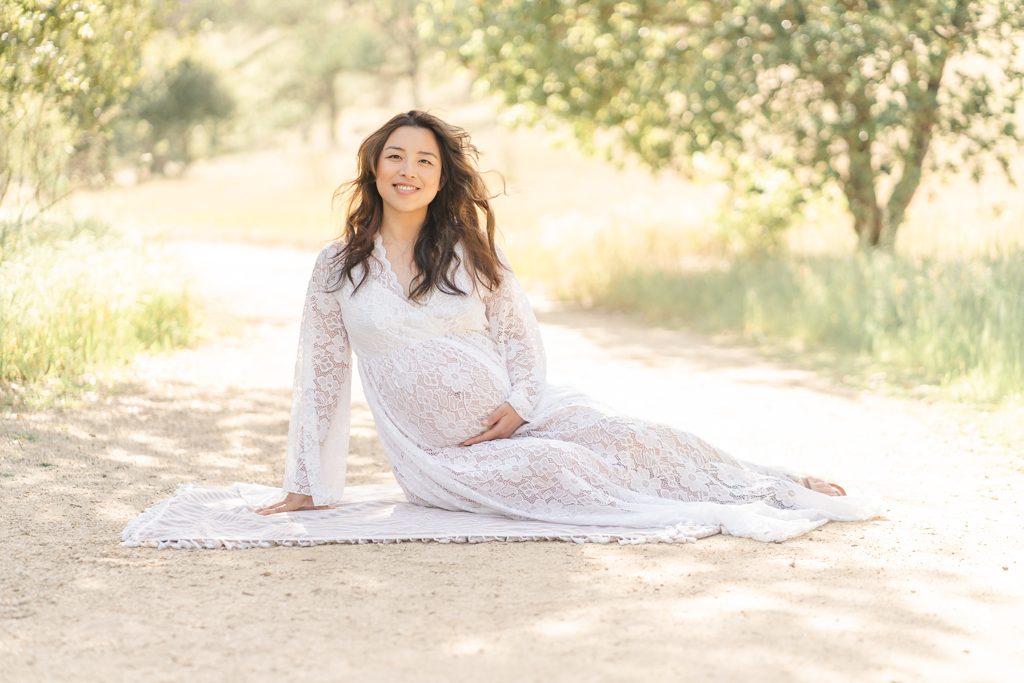 San Jose pregnant woman sitting on the ground at a park wearing a white lacy dress and smiling at the camera