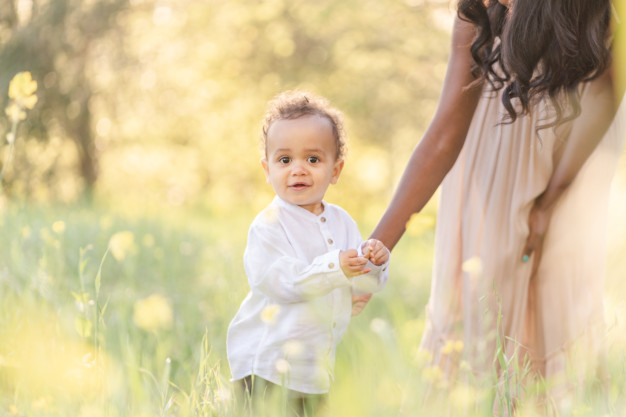 one-year-old boy in a white shirt standing in a field of mustard flowers holding his mommy's hand