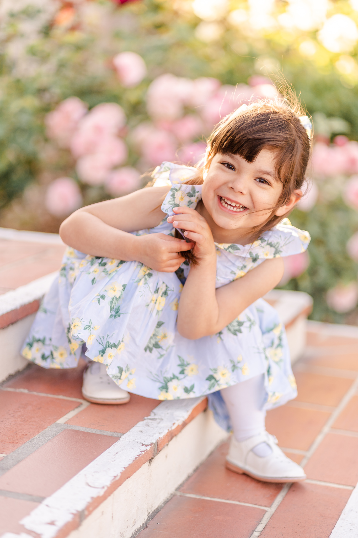 3-year-old girl sitting on steps at the Santa Clara University Mission smiling big with pink roses in the background