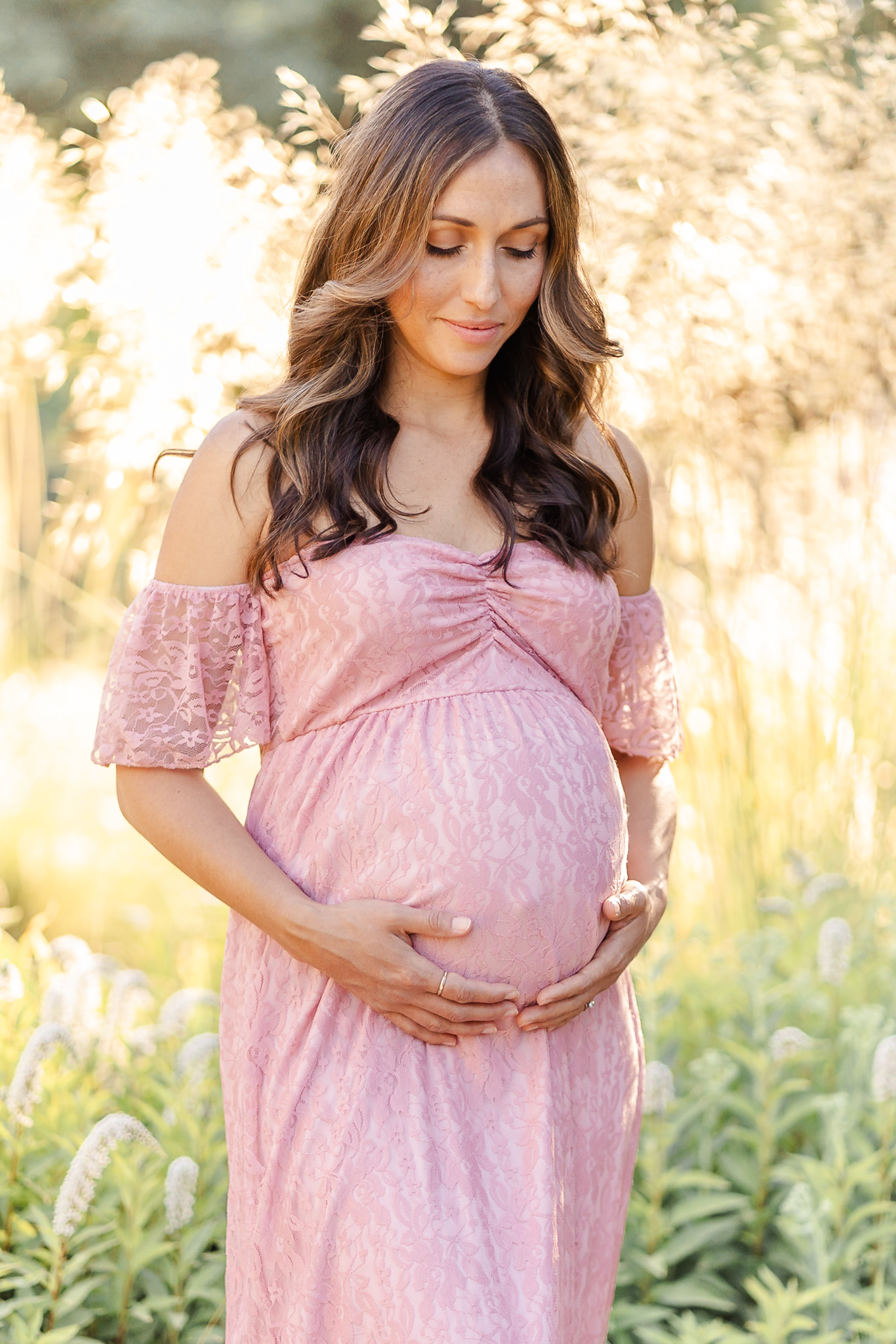 glow of foliage behind a pregnant woman in a pink lacy dress holding her belly and looking down on it.