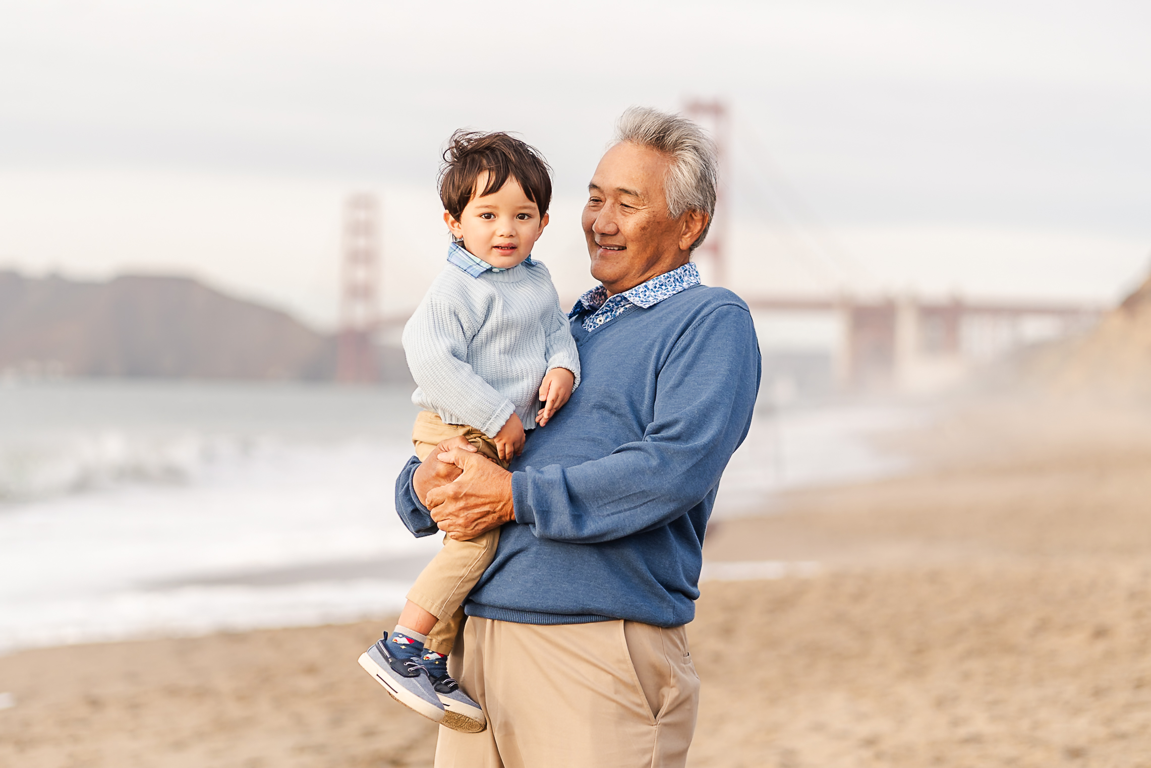 grandpa holding his grandson on Baker Beach with the Golden Gate Bridge in the background